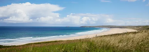 Hayle to Godrevy beach This landscape photograph captures Hayle to Godrevy beach along the coastline of Cornwall, England, in the United Kingdom. The image was taken in the morning during the summer season, as evidenced by the bright sunlight and clear blue sky. The wide sandy beach stretches beside the vibrant blue sea, with gentle waves rolling in towards the shore. The photo also features the distinctive Godrevy headland, a notable landmark, which can be seen jutting out in the distance. Coastal grasses are visible in the foreground, further indicating the natural beauty of this beach area in Cornwall.